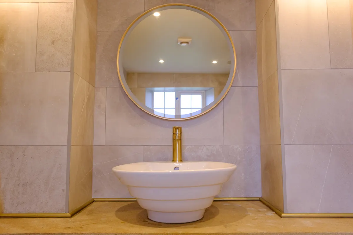 Stone-tiled bathroom with a brass basin tap, brass tile trims and a round mirror by Abbey Group