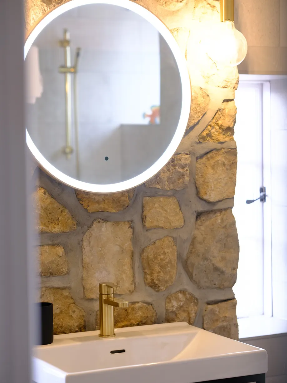 Exposed stone wall, brushed brass tap and a backlit round mirror in a bathroom renovated by Abbey Group